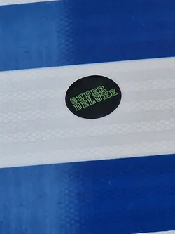 A round black sticker with the text "SUPER DELUXE" in light green.  The sticker is placed on a blue and white striped surface.