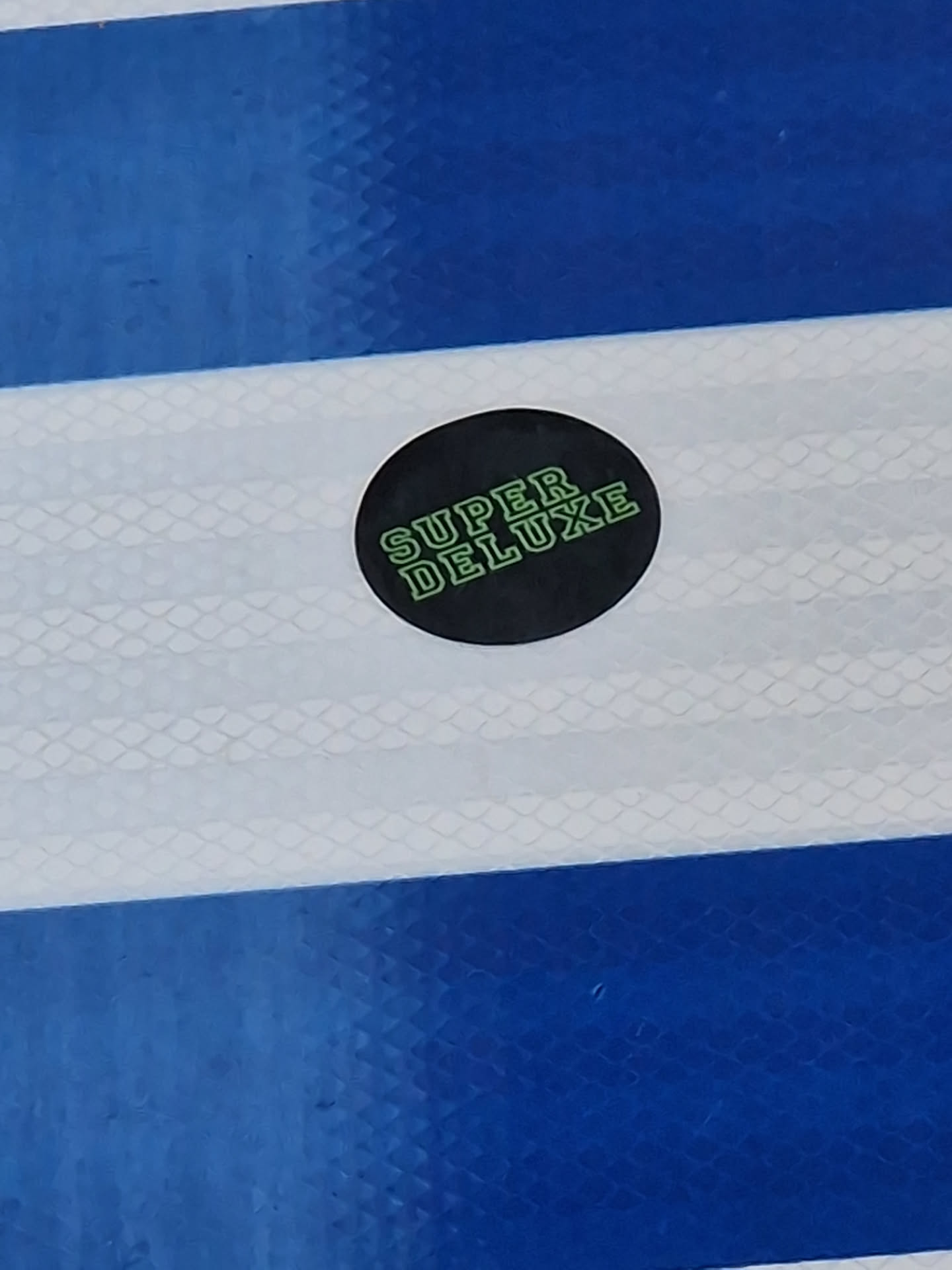 A round black sticker with the text "SUPER DELUXE" in light green.  The sticker is placed on a blue and white striped surface.