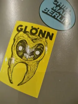 Yellow sticker with a black and white illustration of a grotesque tooth or similar creature. The word GLÖNN is written in black capital letters at the top of the sticker.