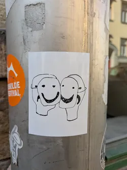 A black and white sticker shows three stylized faces. Each face has a simple line drawing of a happy mask with a wide smile. The sticker is on a metal post.