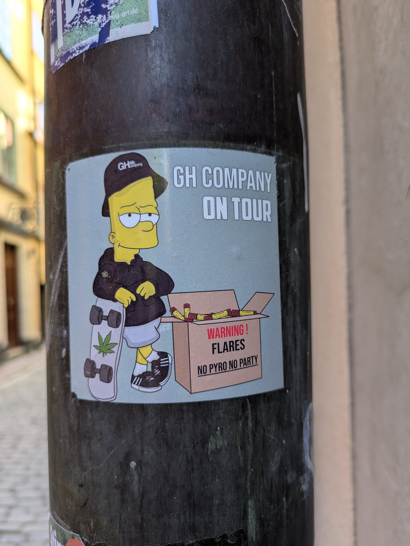 A sticker on a gray pole features Bart Simpson in a black cap and jacket. He stands next to a skateboard with a marijuana leaf design. A box labeled "WARNING! FLARES NO PYRO NO PARTY" is beside him. The text 'GH COMPANY ON TOUR' is above Bart.