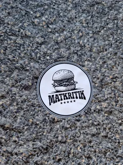 A round sticker featuring a hand-drawn illustration of a burger and the word "MATKRITIK" with four stars underneath.  The style is simple and black and white. The sticker appears to be affixed to a rough textured surface.