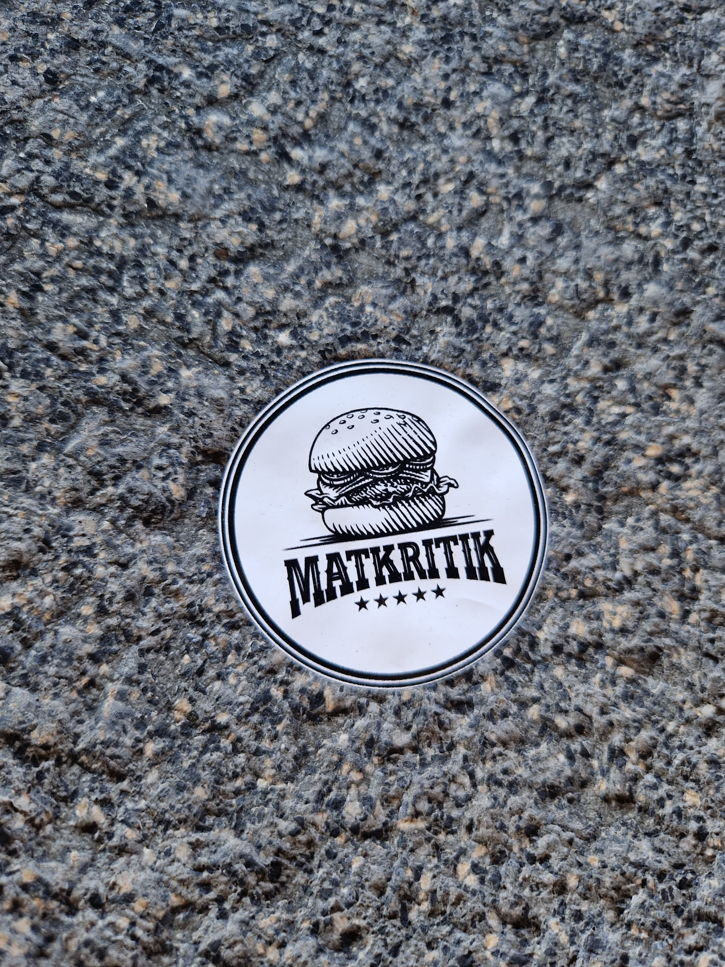 A round sticker featuring a hand-drawn illustration of a burger and the word "MATKRITIK" with four stars underneath.  The style is simple and black and white. The sticker appears to be affixed to a rough textured surface.