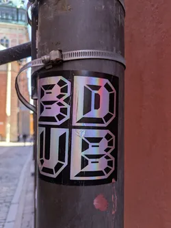 A holographic sticker featuring the text "3D UB" in a stylized, geometric font, affixed to a gray metal pole secured with a metal clamp. The iridescent sheen gives the sticker a dynamic, eye-catching effect.