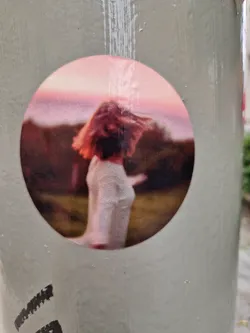 Round sticker featuring a photograph of a woman with short hair walking outdoors at sunset. The colors are muted and the style is reminiscent of film photography. 