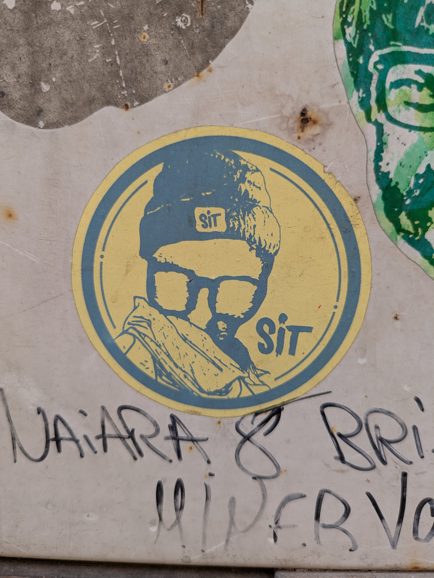This sticker features a stylized, monochromatic portrait of a person wearing glasses and a beanie. The image is primarily blue and yellow, with the person facing forward, giving a direct, focused appearance. The sticker is round and shows some signs of wear, adding to its vintage or street art feel. The word "Sit" is written in a bold, sans-serif font both on the hat and at the bottom of the sticker.