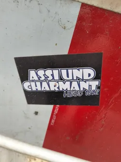 A black sticker with white and blue text that says "ASSLUND CHARMANT" and "#HIBSC 1892" in a stylized font. The sticker is placed on a white and red surface.