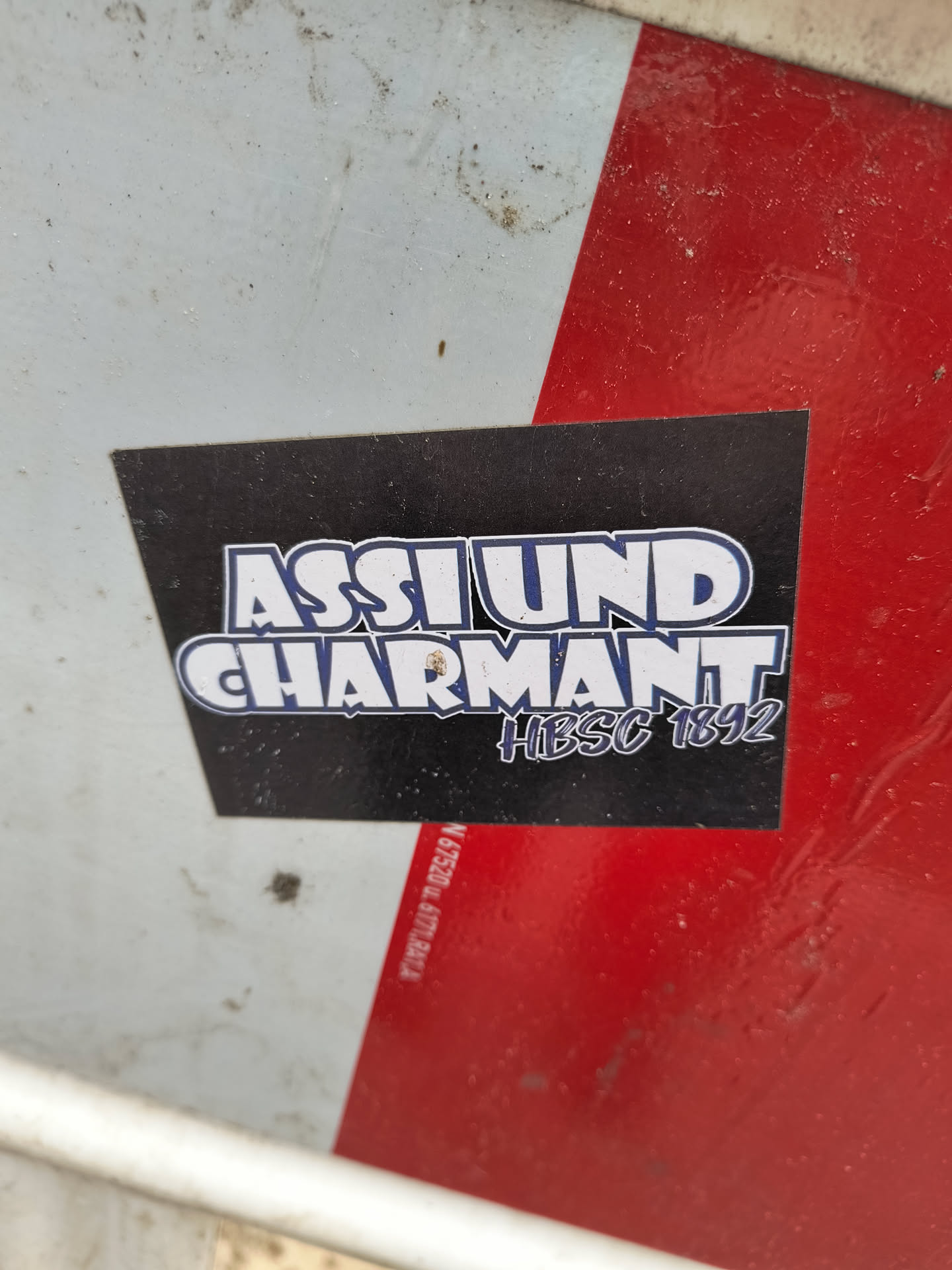 A black sticker with white and blue text that says "ASSLUND CHARMANT" and "#HIBSC 1892" in a stylized font. The sticker is placed on a white and red surface.