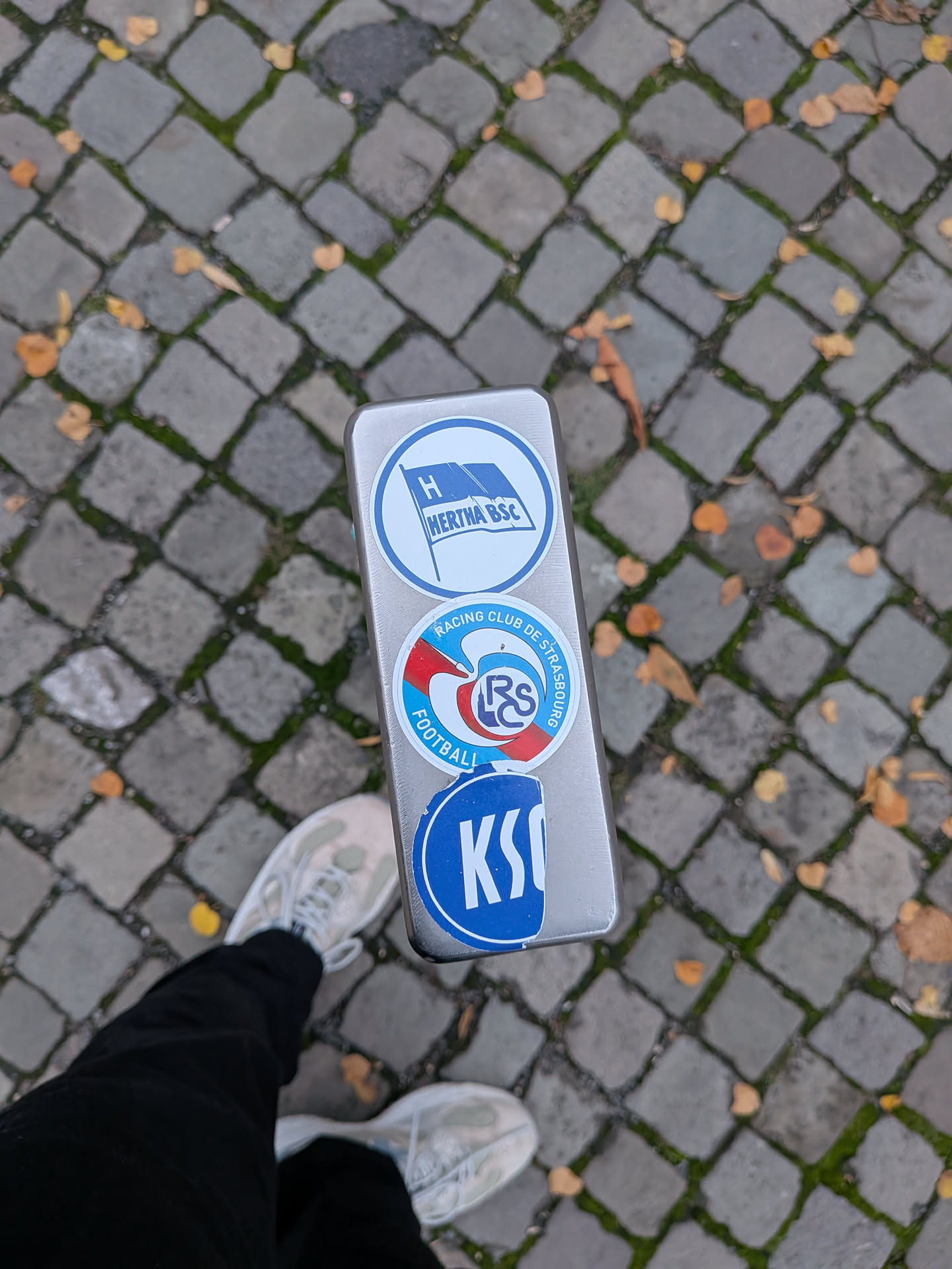 A circular sticker displaying the crest of the French football team Racing Club de Strasbourg. The logo features a light blue circular border inscribed with 'RACING CLUB DE STRASBOURG FOOTBALL'. The central design incorporates red, white, and blue sweeping shapes around an inner circle containing the stylized interlocking club monogram 'RCS'. It is affixed to the metallic surface of a post between two other football club stickers.