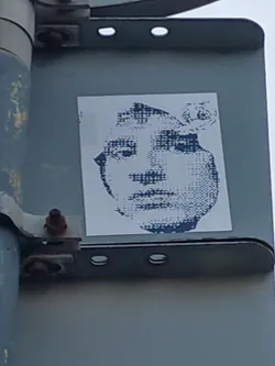 A sticker featuring a halftone portrait of a person's face. The style is reminiscent of pop art and printmaking techniques. The sticker is affixed to a metal pole.