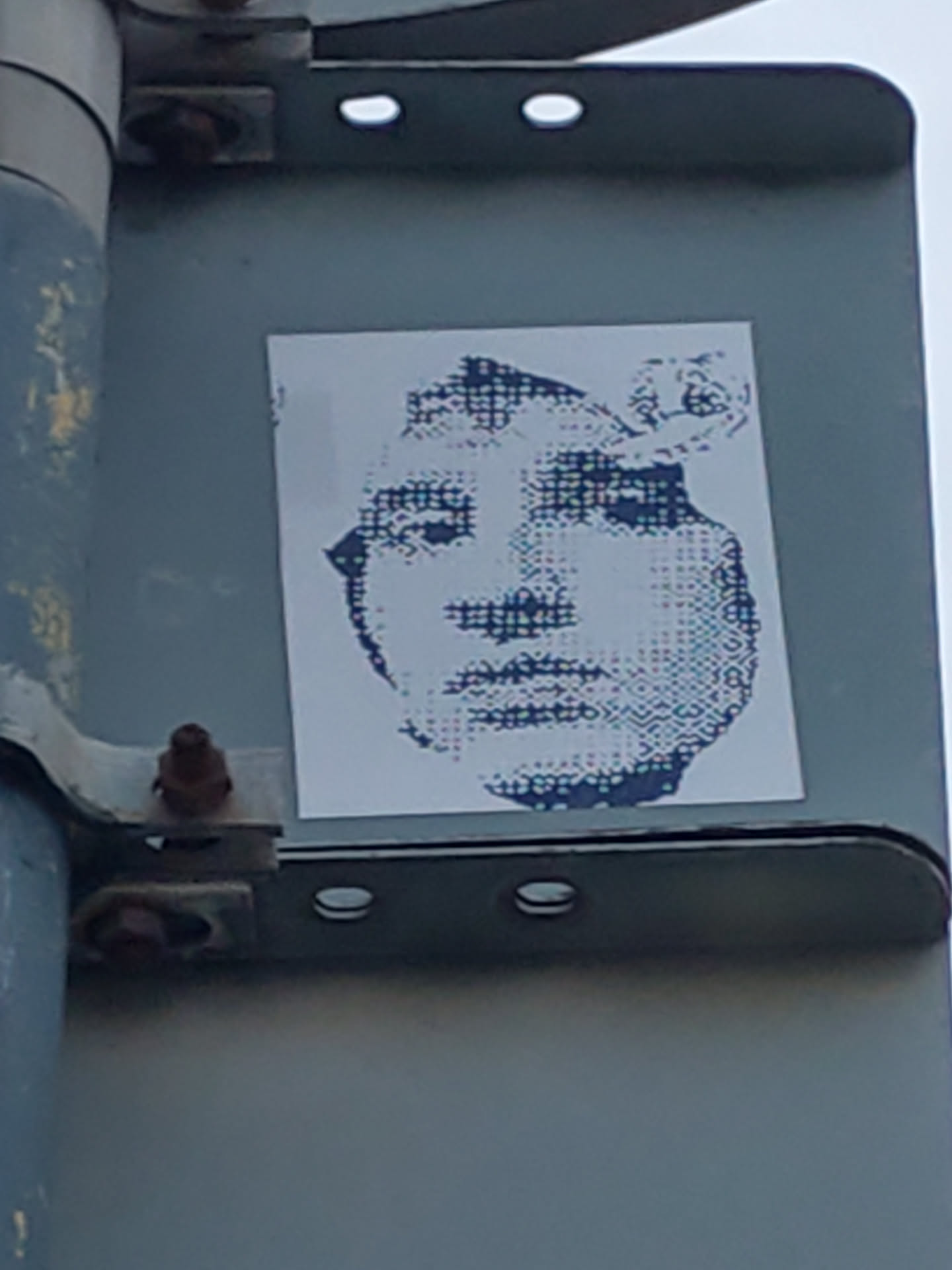 A sticker featuring a halftone portrait of a person's face. The style is reminiscent of pop art and printmaking techniques. The sticker is affixed to a metal pole.