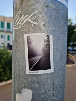 A black and white photograph of a foggy road. The sticker is attached to a metal pole. The Instagram handle @drzdowcz is visible at the bottom of the sticker.