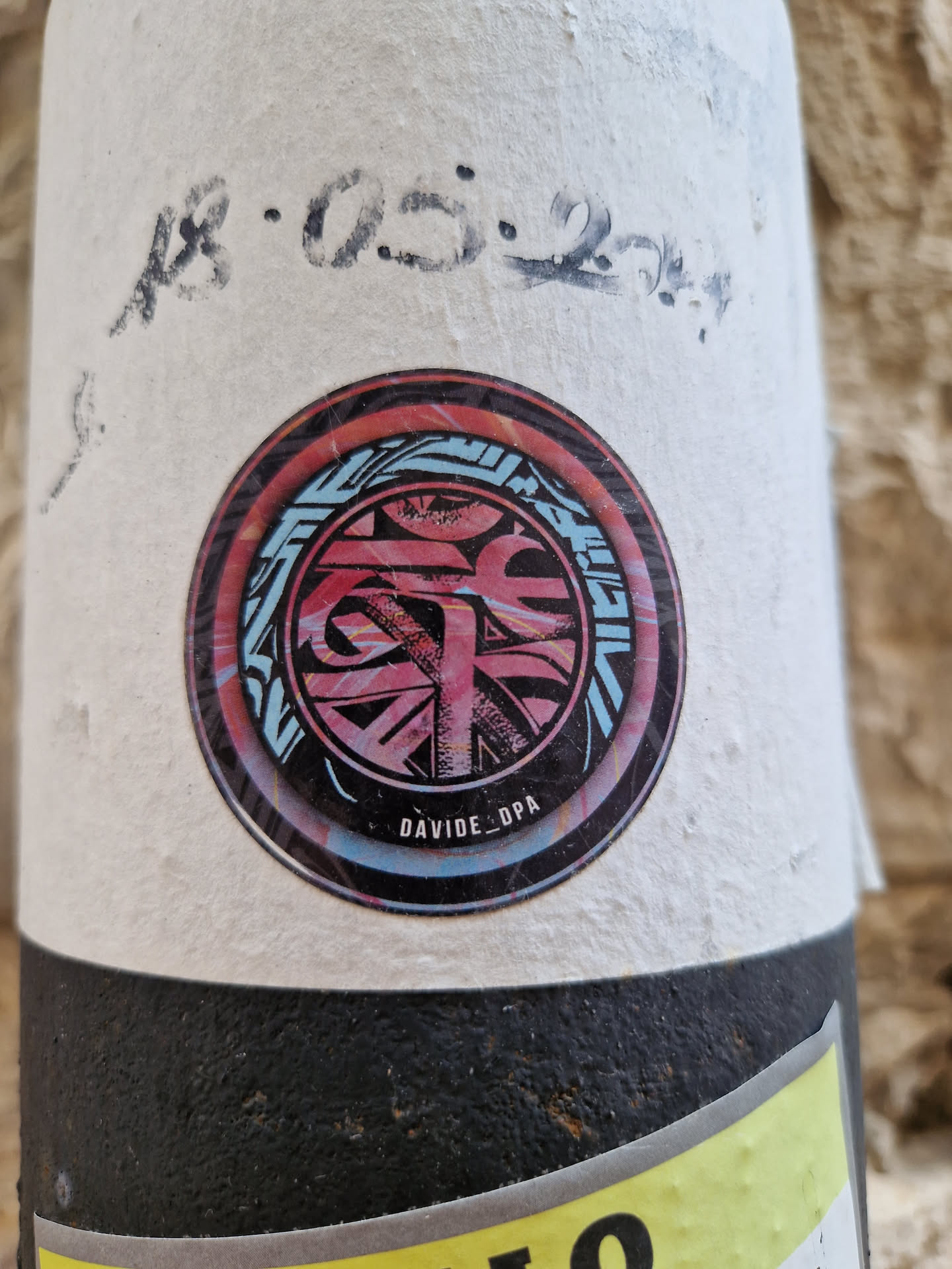 A round sticker featuring an abstract design in shades of pink, blue, and black. The design is circular and contains various shapes and lines, creating a visually appealing pattern. At the bottom of the sticker, the artist's signature \"DAVIDE_DPA\" is written.