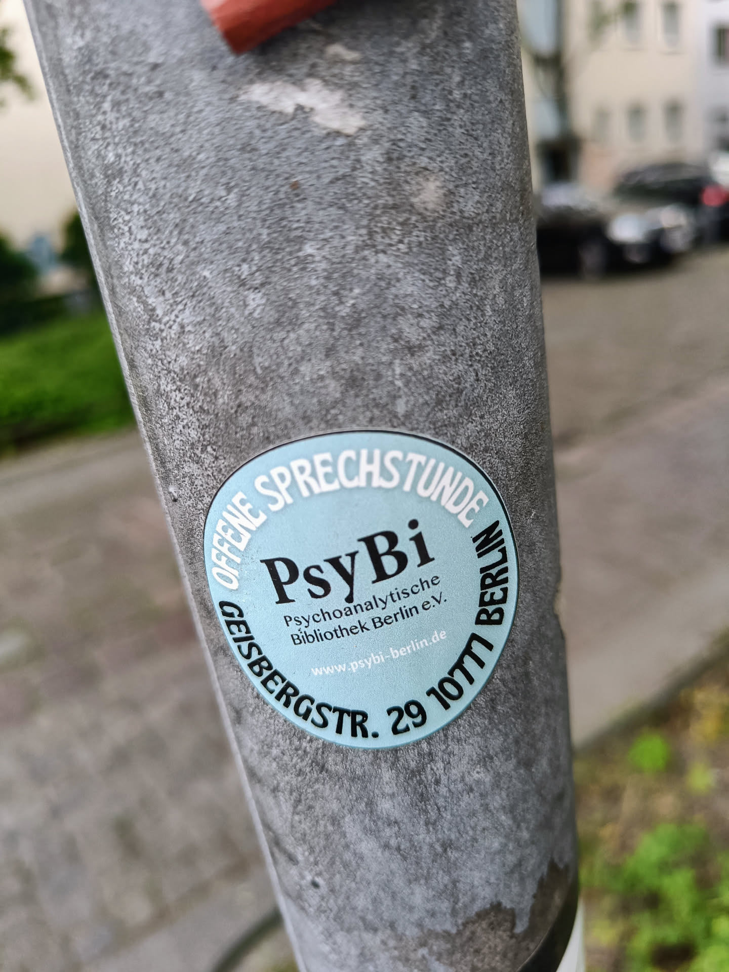 A round, light blue sticker with the text "PsyBi" prominently displayed.  Smaller text indicates it's for the Psychoanalytic Library in Berlin, including their address and website.  The sticker is affixed to a grey metal pole.