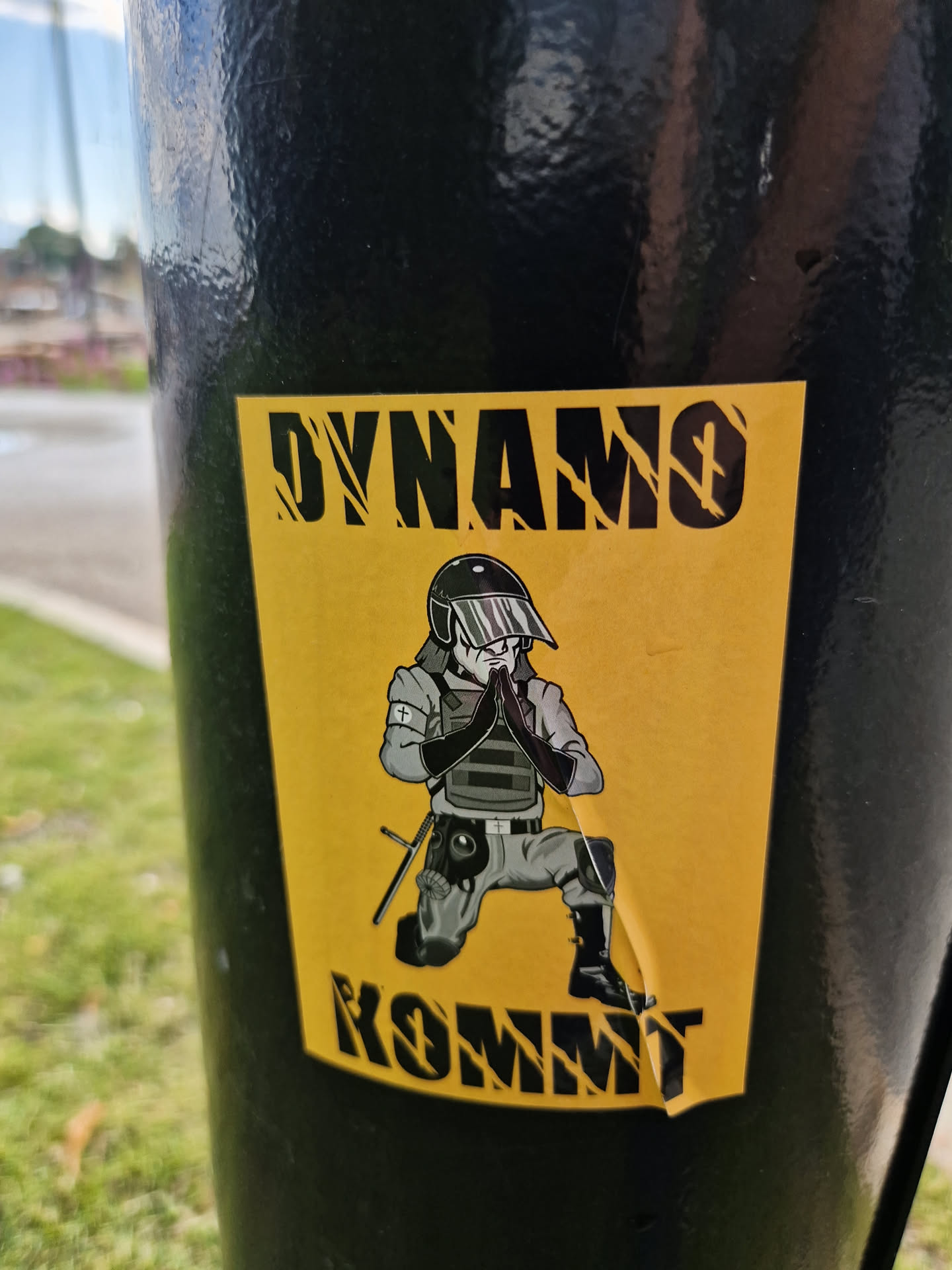 A yellow sticker with a black illustration of a police officer kneeling with hands together in prayer. The words "DYNAMO" and "KOMMT" are written in black stylized font at the top and bottom of the sticker respectively.