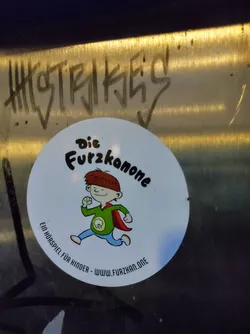 A round sticker featuring a cartoon illustration of a child superhero running. The text on the sticker says: "Die Furzkanone" which translates to "The Fart Cannon" in English, followed by a tagline in German stating it's an audio play for children. A website address is also visible.