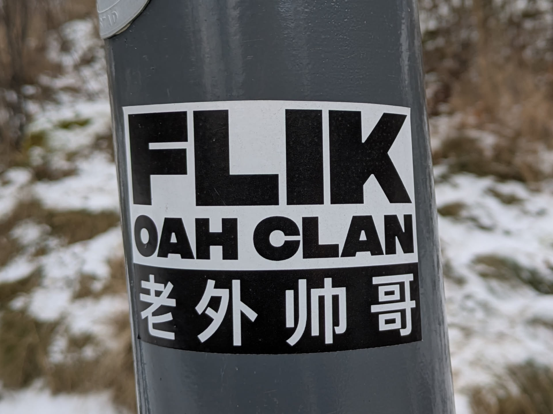 The sticker is adhered to a gray metal pole. The sticker is white with black text reading "FLIK OAH CLAN". Below that there are two lines with chinese characters.