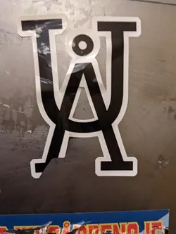 A close-up shot of a sticker featuring a stylized logo composed of interlocking uppercase letters T and U, creating a visually striking emblem. The design is minimalist, using only black and white. The sticker appears to be affixed to a metallic surface.