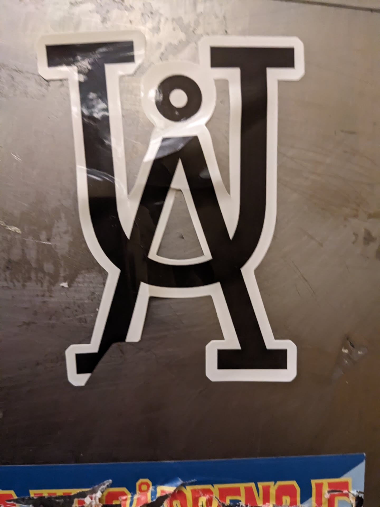 A close-up shot of a sticker featuring a stylized logo composed of interlocking uppercase letters T and U, creating a visually striking emblem. The design is minimalist, using only black and white. The sticker appears to be affixed to a metallic surface.