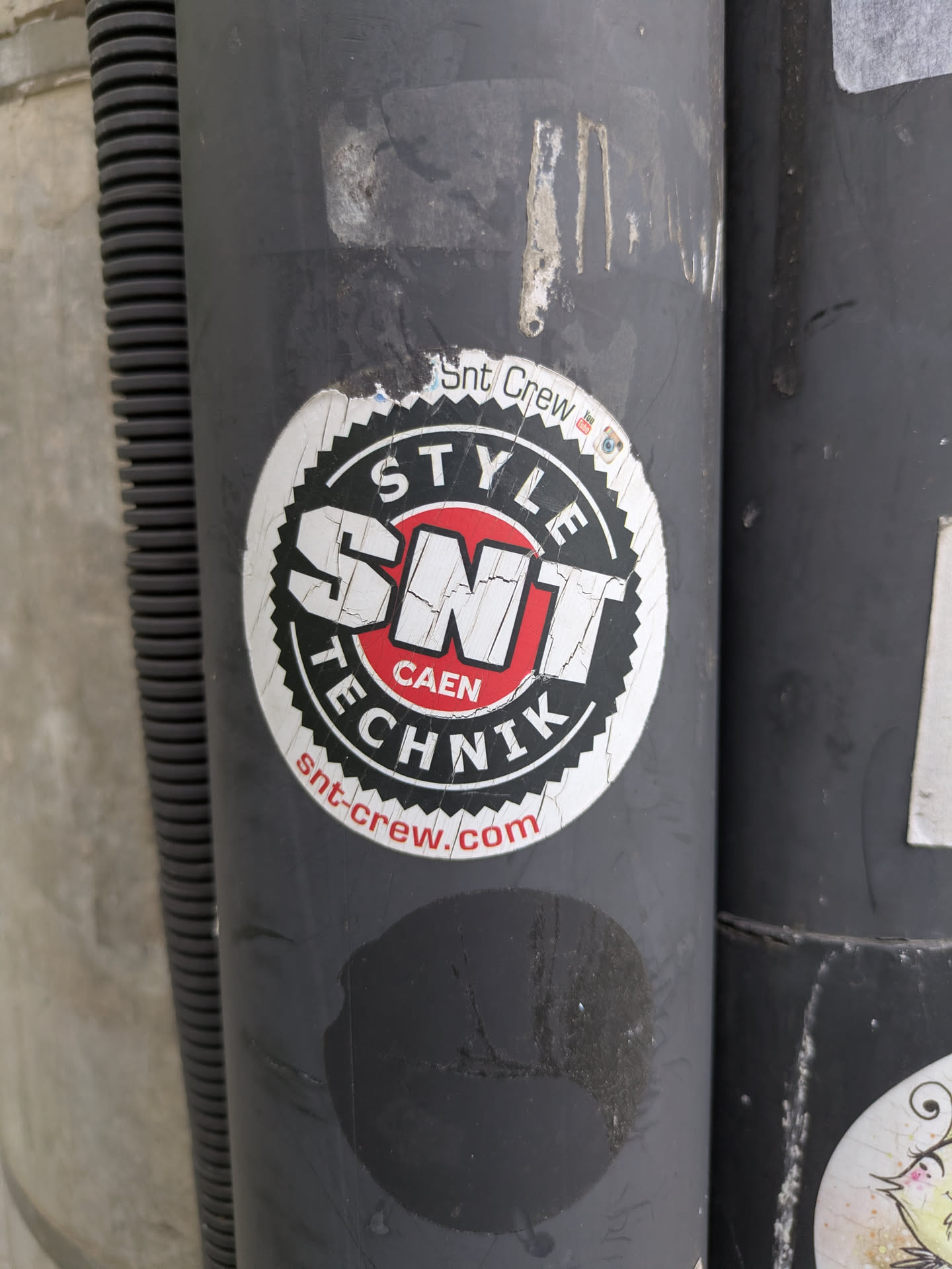 A round, slightly damaged sticker is affixed to a gray, cylindrical surface. The sticker features the text 'SNT Crew', 'STYLE', 'SNT CAEN', 'TECHNIK', and 'snt-crew.com'. The logo is designed in a black and white color scheme with red accents. The sticker appears to be exposed to the elements and shows signs of wear and tear.