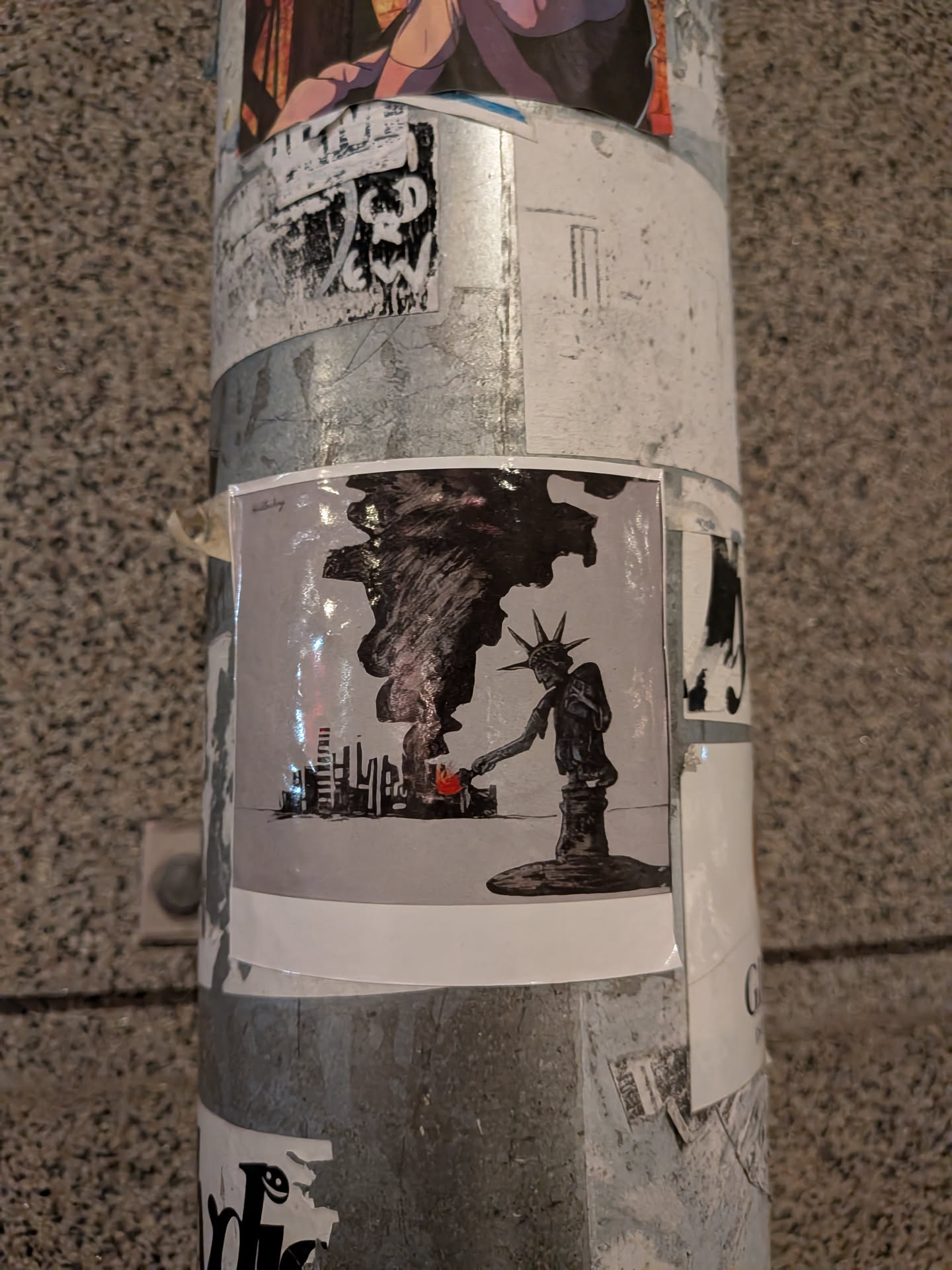 The sticker depicts a stylized, dark silhouette of the Statue of Liberty, appearing to be made of stone or metal. She is shown reaching down with her right hand, holding a small, bright red flame towards a miniature cityscape. The cityscape is rendered in black and white with sharp, angular buildings. Above the city and the statue, a large, dark, swirling mass resembling a tornado or a storm cloud dominates the upper portion of the sticker. The background is a light gray, and the overall impression is one of impending doom or destruction.
