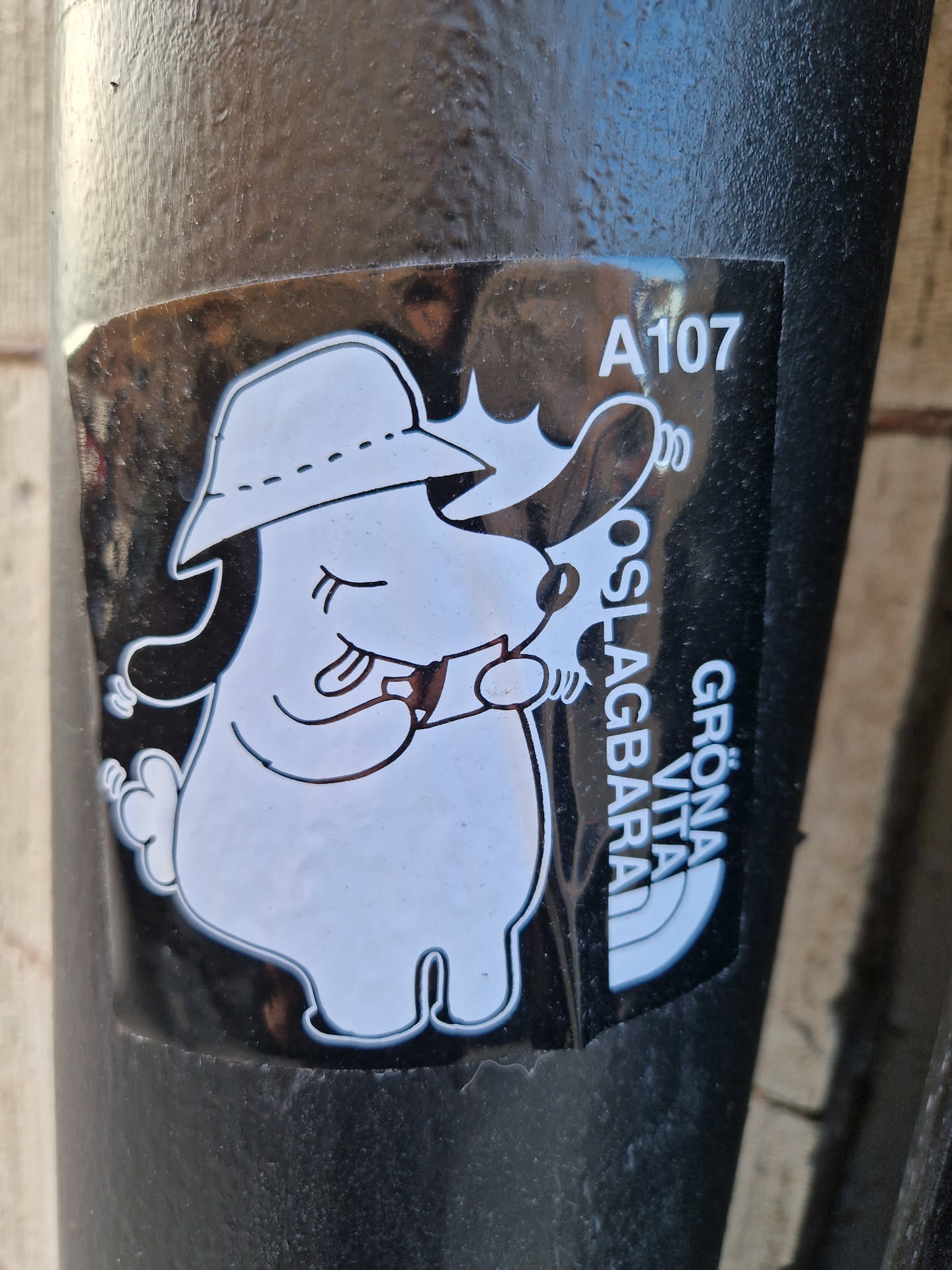 A black and white sticker featuring a cartoon illustration of a dog-like character wearing a hat.  The character appears to be holding or looking at something. The text "A107", "OSLAGBARA", and "GRÖNA VITA" is visible on the sticker, likely referring to a product, brand or location. The style is minimalist and cute.