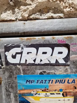 A black and white sticker with the word "GRRR" in a bold, uppercase font. The sticker is affixed to a weathered metal surface. Another sticker is visible underneath, featuring Italian text and an image of a boat. The overall aesthetic suggests urban street art or graffiti.