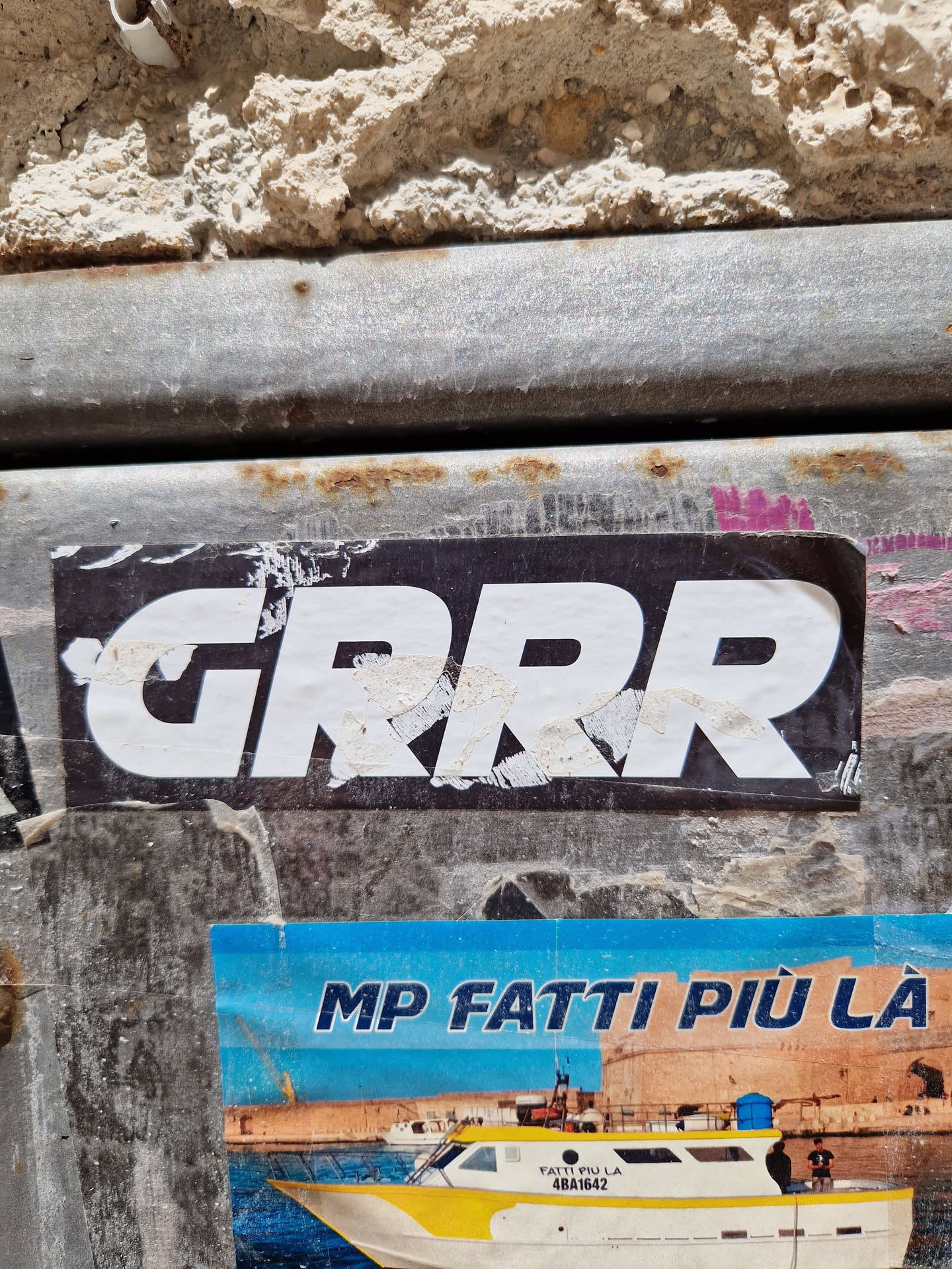 A black and white sticker with the word "GRRR" in a bold, uppercase font. The sticker is affixed to a weathered metal surface. Another sticker is visible underneath, featuring Italian text and an image of a boat. The overall aesthetic suggests urban street art or graffiti.
