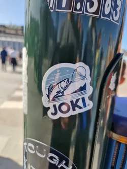 A sticker featuring a fish jumping out of the water with the brand name Joki. The sticker has a minimalist design with a blue, green and white color scheme.