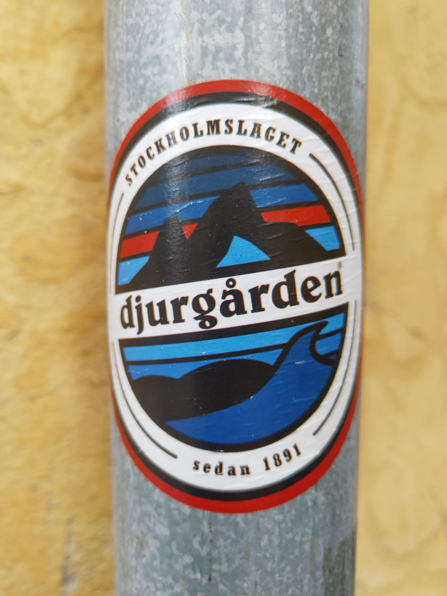 A round sticker featuring a stylized landscape of Djurgården, Stockholm. The design includes mountains, water, and a wave, with the text "Stockholmslaget" at the top and "Djurgården" prominently in the center.  "Sedan 1891" is written at the bottom, indicating the year of establishment.