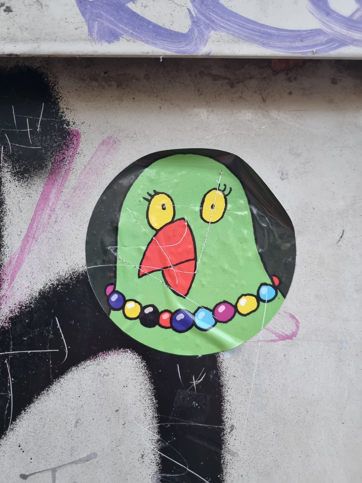 This charming sticker features a cartoon bird with bright yellow eyes, a red beak, and a colorful beaded necklace. The bird's expression is sweet and slightly surprised. The sticker is round and has a slightly worn look, which adds to its character. It's been affixed to a surface with some existing graffiti, creating a nice contrast between the cute sticker and the urban backdrop.
