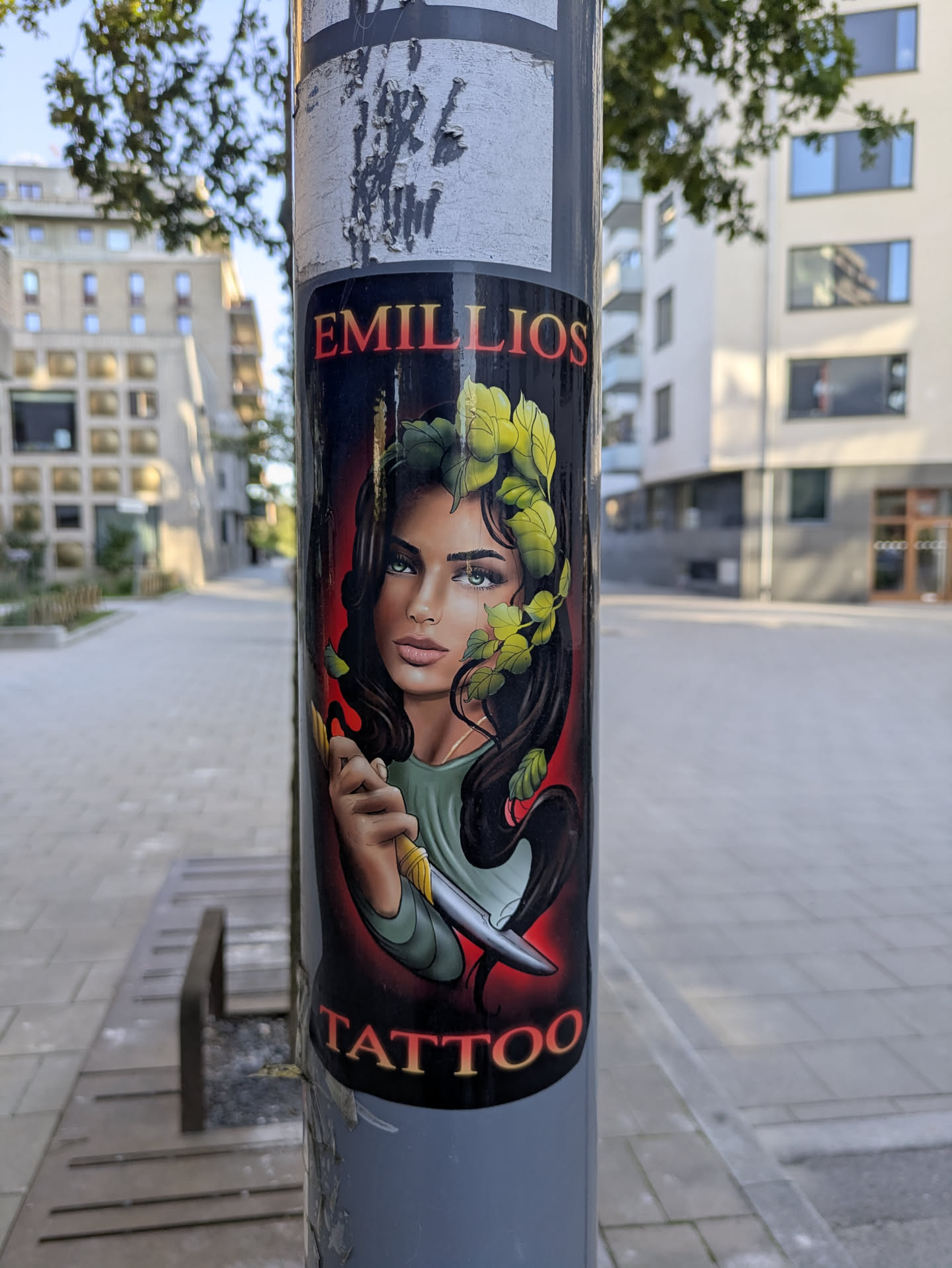 The sticker prominently displays a stylized portrait of a woman with piercing blue eyes, dark wavy hair intertwined with vibrant green leaves, and delicate facial features. She is depicted holding a gleaming, sharp knife. The background transitions from a fiery red glow behind her head to black. The text 'EMILLIOS' is arched above her head in bold, red lettering with a yellow outline, and 'TATTOO' is displayed below her chin in similar, but slightly smaller, yellow lettering with a red outline. The overall style evokes a traditional tattoo aesthetic.