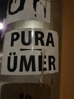 A black and white sticker with the text "PURA ÜMER" on it. The sticker is attached to a metal pole.