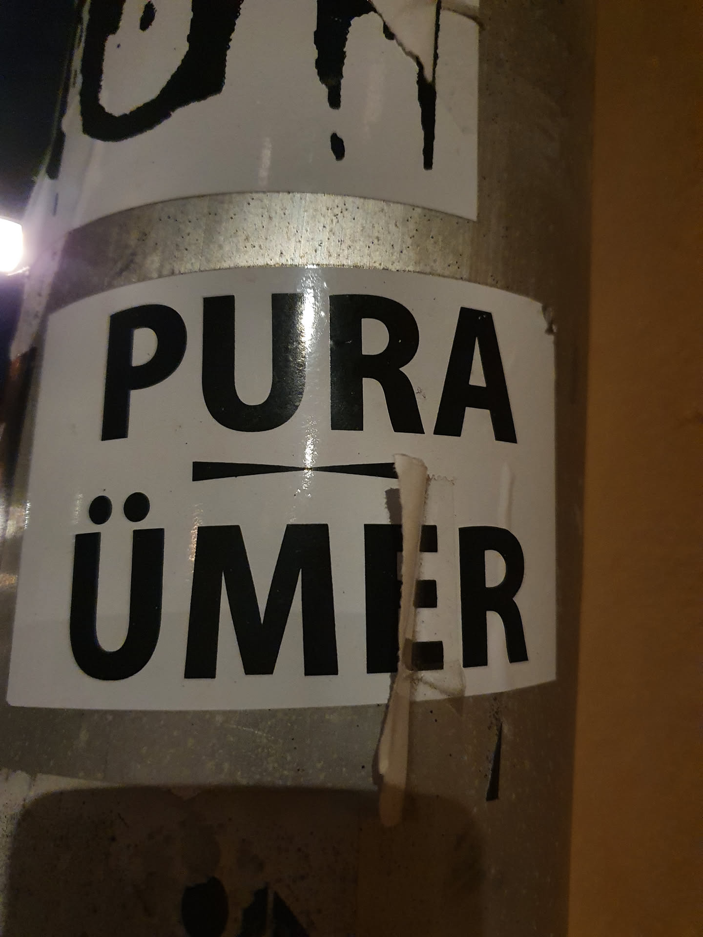A black and white sticker with the text "PURA ÜMER" on it. The sticker is attached to a metal pole.