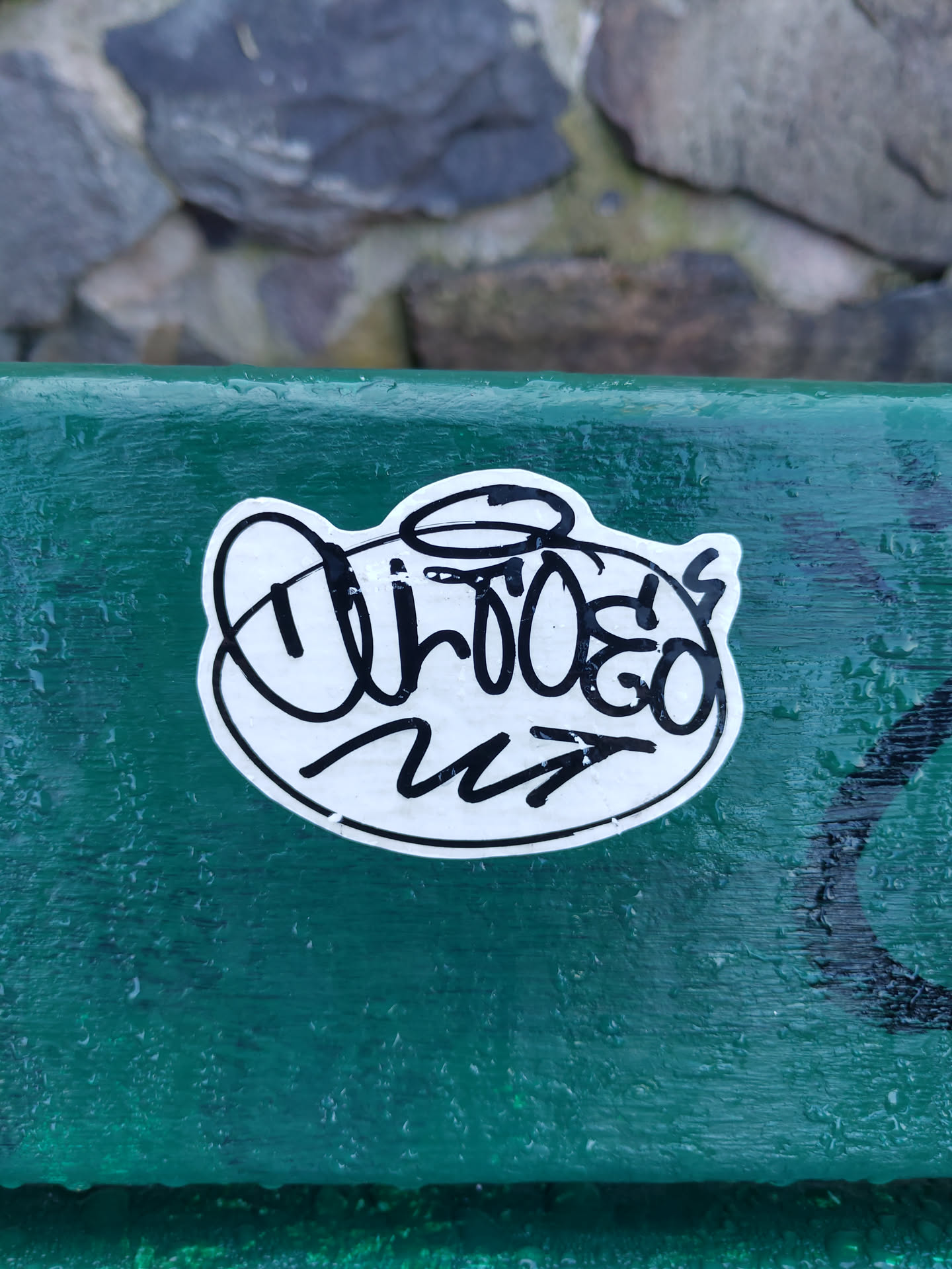 A white oval sticker with black ink drawing and text. The main text says "Urboego" with an arrow pointing right. The drawing looks like a stylized figure with a halo.