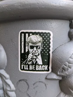 A black and white sticker featuring an illustration of Donald Trump, wearing sunglasses and pointing directly forward, is adhered to a textured gray surface. The backdrop behind Trump features the design of the American flag. Below Trump, the phrase "I'LL BE BACK" is printed in bold capital letters. The sticker has a border.