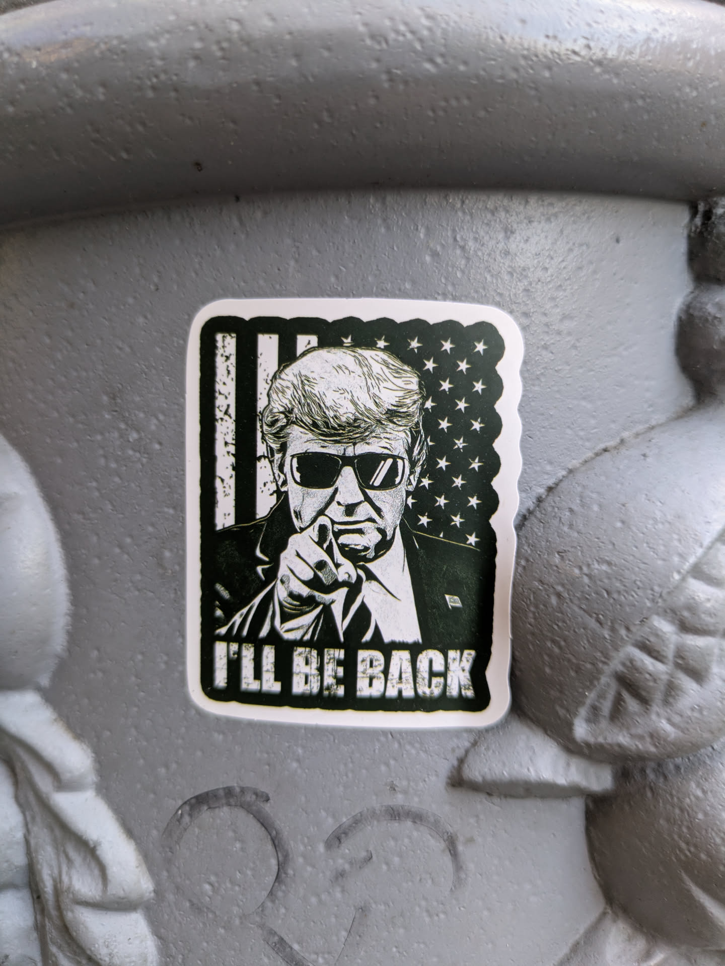 A black and white sticker featuring an illustration of Donald Trump, wearing sunglasses and pointing directly forward, is adhered to a textured gray surface. The backdrop behind Trump features the design of the American flag. Below Trump, the phrase "I'LL BE BACK" is printed in bold capital letters. The sticker has a border.