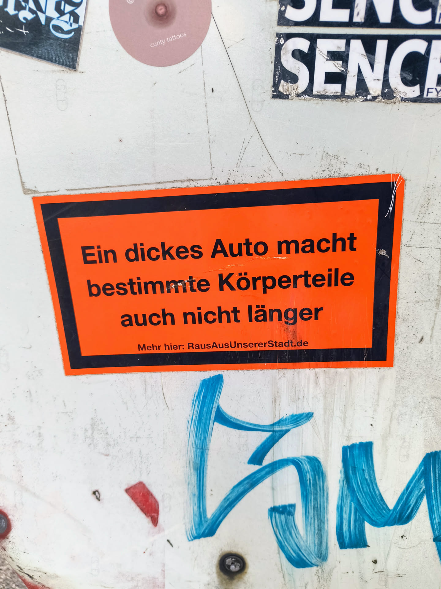 The sticker is orange with black text and a black border. It says: "Ein dickes Auto macht bestimmte Körperteile auch nicht länger. Mehr hier: RausAusUnsererStadt.de" which translates to "A big car doesn't make certain body parts longer either. More here: RausAusUnsererStadt.de".