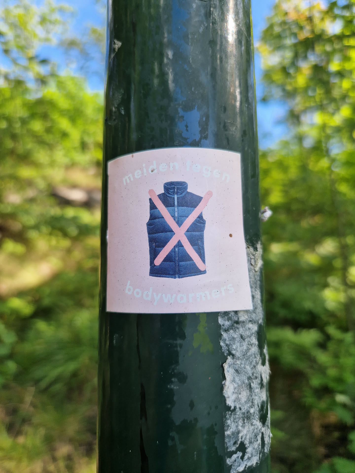 A sticker with a crossed-out image of a body warmer and the text "meiden tegen bodywarmers" which translates from Dutch to "avoid body warmers".