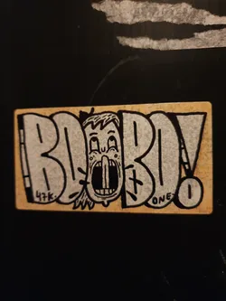 A beige sticker with black ink features a cartoon character's face with an open mouth, seemingly screaming, placed centrally within the stylized lettering spelling out "BOBOLO."  The style is reminiscent of graffiti or street art.  There are small details, including "47k." and "ONE" written in the art. A small number '1' is visible in a circle. 