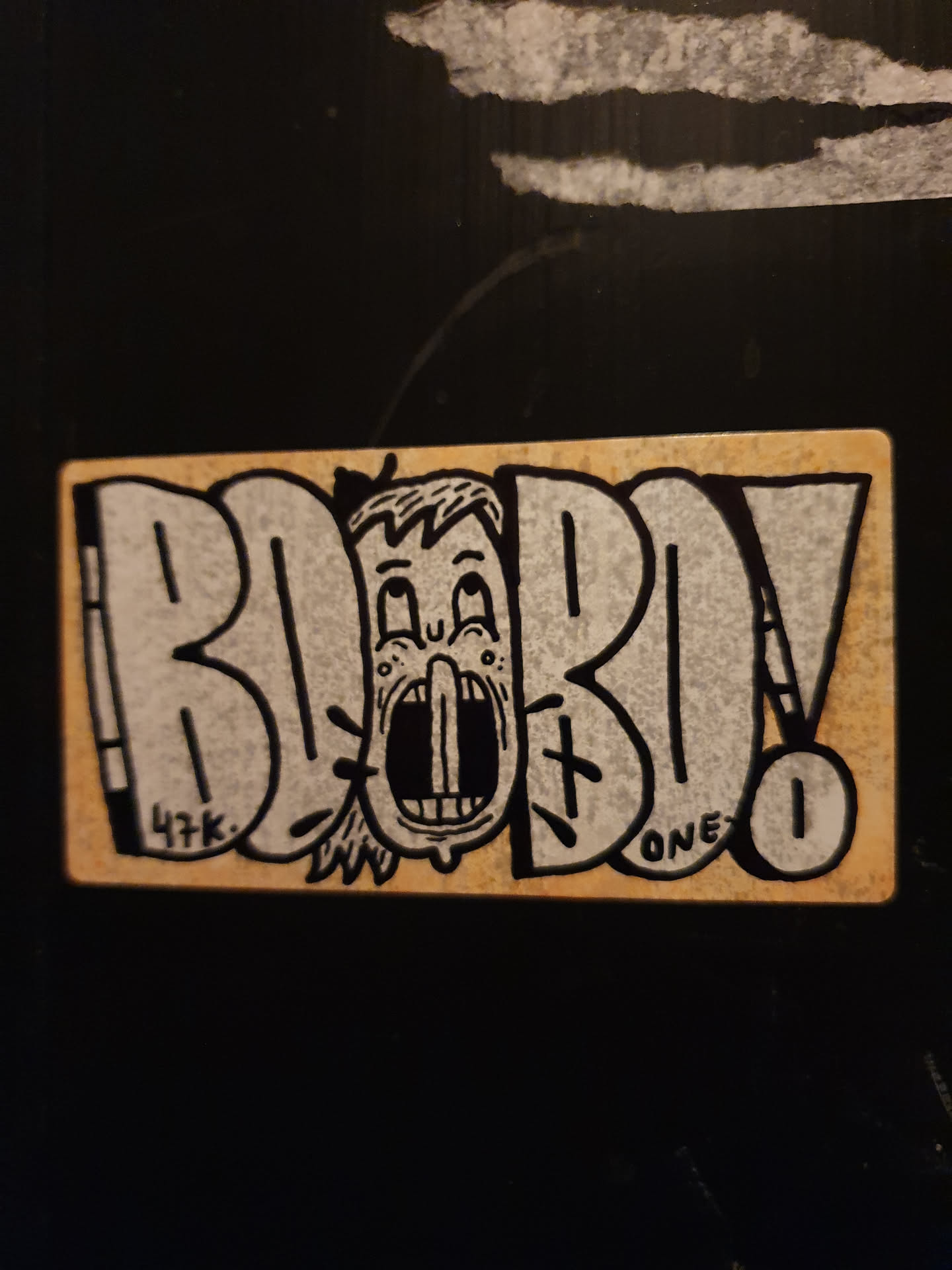 A beige sticker with black ink features a cartoon character's face with an open mouth, seemingly screaming, placed centrally within the stylized lettering spelling out "BOBOLO."  The style is reminiscent of graffiti or street art.  There are small details, including "47k." and "ONE" written in the art. A small number '1' is visible in a circle. 