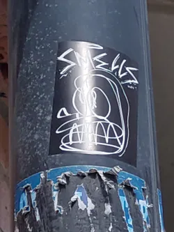 A black and white sticker with a cartoonish face and the word "Smells" written in a stylized font. The sticker is on a dark grey pole.  The sticker has a slightly distressed look.