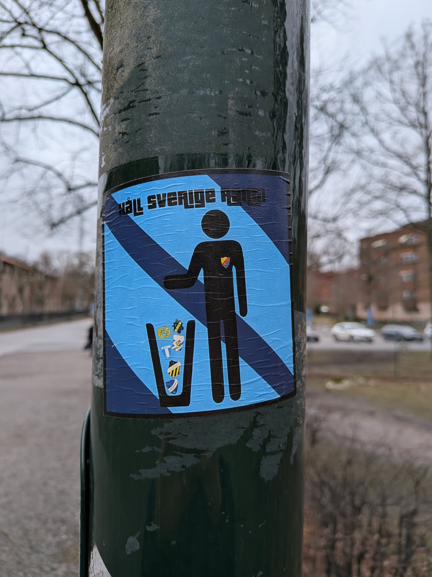 The sticker is rectangular, featuring a light blue background with darker blue diagonal stripes. A black silhouette of a person is depicted in the center, appearing to drop various colorful football club crests into a black trash can. The person's chest bears a small, colorful crest, identifiable as Djurgårdens IF's logo. At the top of the sticker, black block letters read 'HÅLL SVERIGE RENT!'. The sticker shows signs of wear, with visible wrinkles and slight peeling, adhered to a dark green pole.