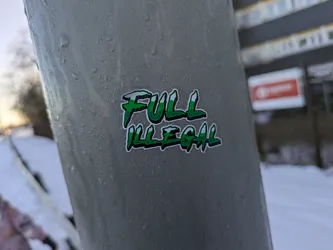 A green and white sticker with the words "FULL ILLEGAL" written in a stylized font. The sticker is affixed to a gray metal surface outdoors, possibly a pole or pipe.  The background is blurred but shows a snowy scene.