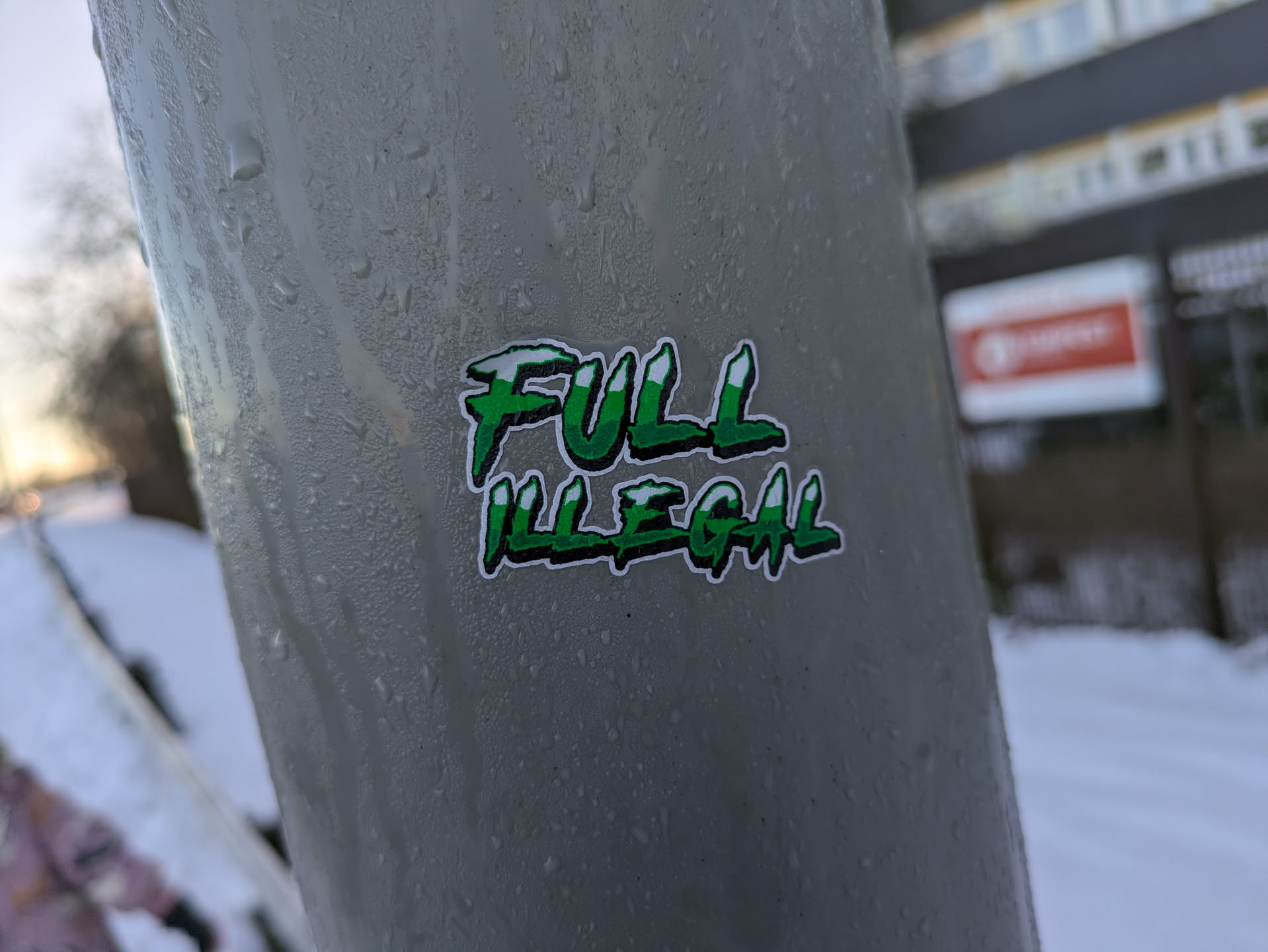 A green and white sticker with the words "FULL ILLEGAL" written in a stylized font. The sticker is affixed to a gray metal surface outdoors, possibly a pole or pipe.  The background is blurred but shows a snowy scene.