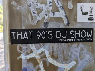 The central sticker is a rectangular black label with prominent white text. The main text reads "THAT 90'S DJ SHOW" in a rounded, somewhat distressed font, suggesting a retro or casual style. Below this, smaller text indicates "INSTAGRAM: @THAT90S_SHOW". The sticker is applied to a textured, light brown or beige surface, possibly a utility box, which shows signs of other white and grey paint marks or graffiti. To the top right, another small white sticker depicts a black and white image of a man holding a trophy.