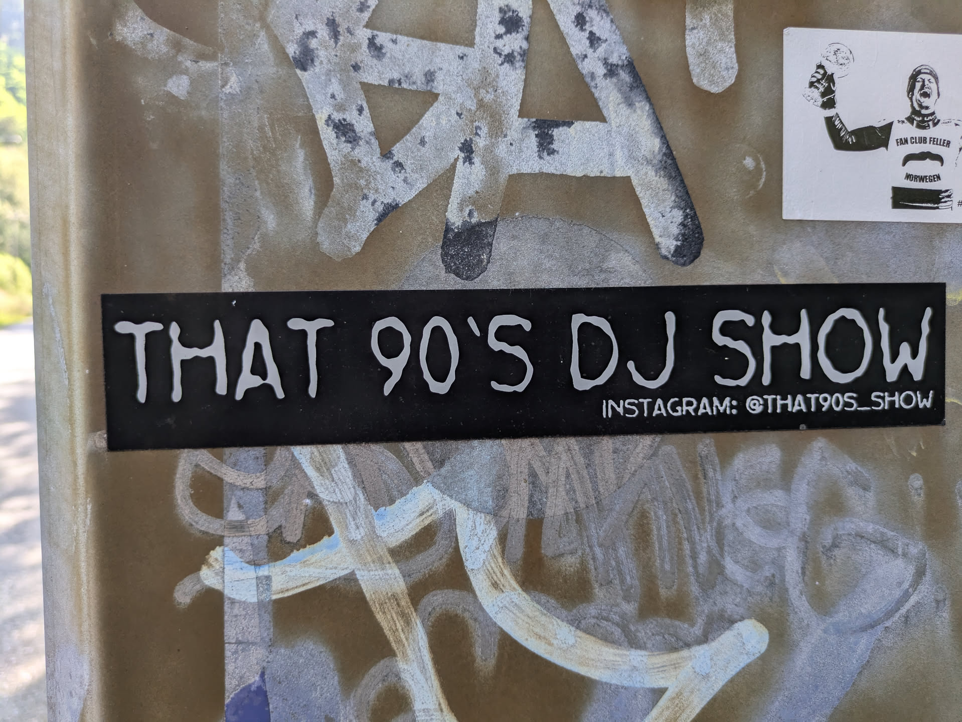 The central sticker is a rectangular black label with prominent white text. The main text reads "THAT 90'S DJ SHOW" in a rounded, somewhat distressed font, suggesting a retro or casual style. Below this, smaller text indicates "INSTAGRAM: @THAT90S_SHOW". The sticker is applied to a textured, light brown or beige surface, possibly a utility box, which shows signs of other white and grey paint marks or graffiti. To the top right, another small white sticker depicts a black and white image of a man holding a trophy.