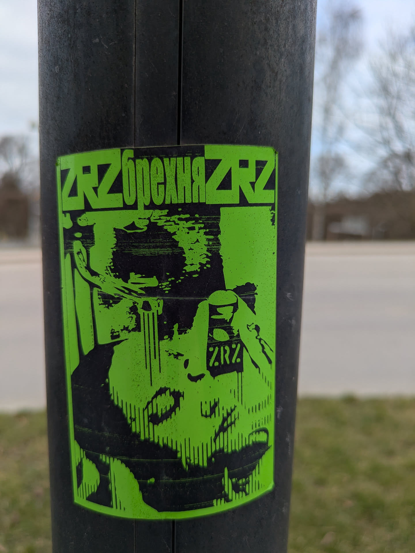 A sticker on a black pole featuring an abstract image in black and green. The word "ZrzbrexняZrz" is printed on top of the image. The image appears to depict a person and some kind of equipment, possibly a medical or scientific device.