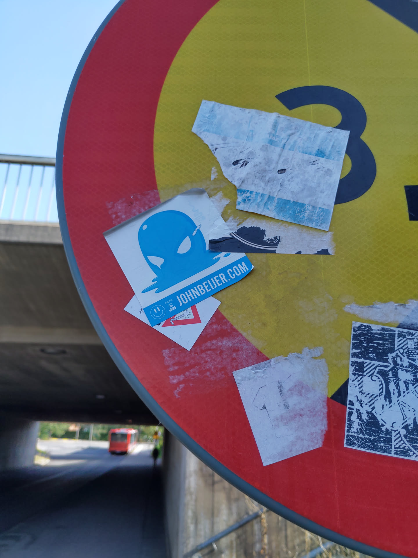 A blue alien head sticker on a traffic sign. The sticker has the website johnbeijer.com on it. There are other stickers on the sign too.