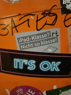 A sticker with the text "IT'S OK" in neon blue letters on a black background.  Above it is a smaller sticker in German that says "iPad-Klasse? Nicht so klasse!" which translates to "iPad-class? Not so classy!"